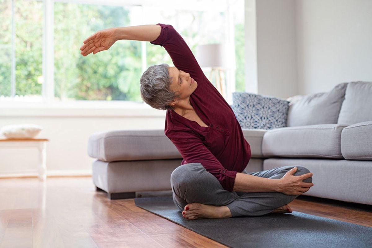 Older woman sitting cross-legged on a yoga mat at home, reaching one arm overhead in a seated side stretch.