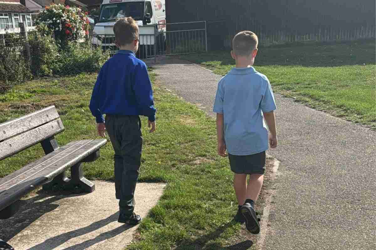 Two young boys walk side by side on a sunny day, heading down a paved path near a park bench. One wears a blue school uniform with long pants, and the other wears a light blue polo shirt with shorts. Their backs are to the camera as they stroll toward a residential area, creating a quiet, everyday moment of childhood.