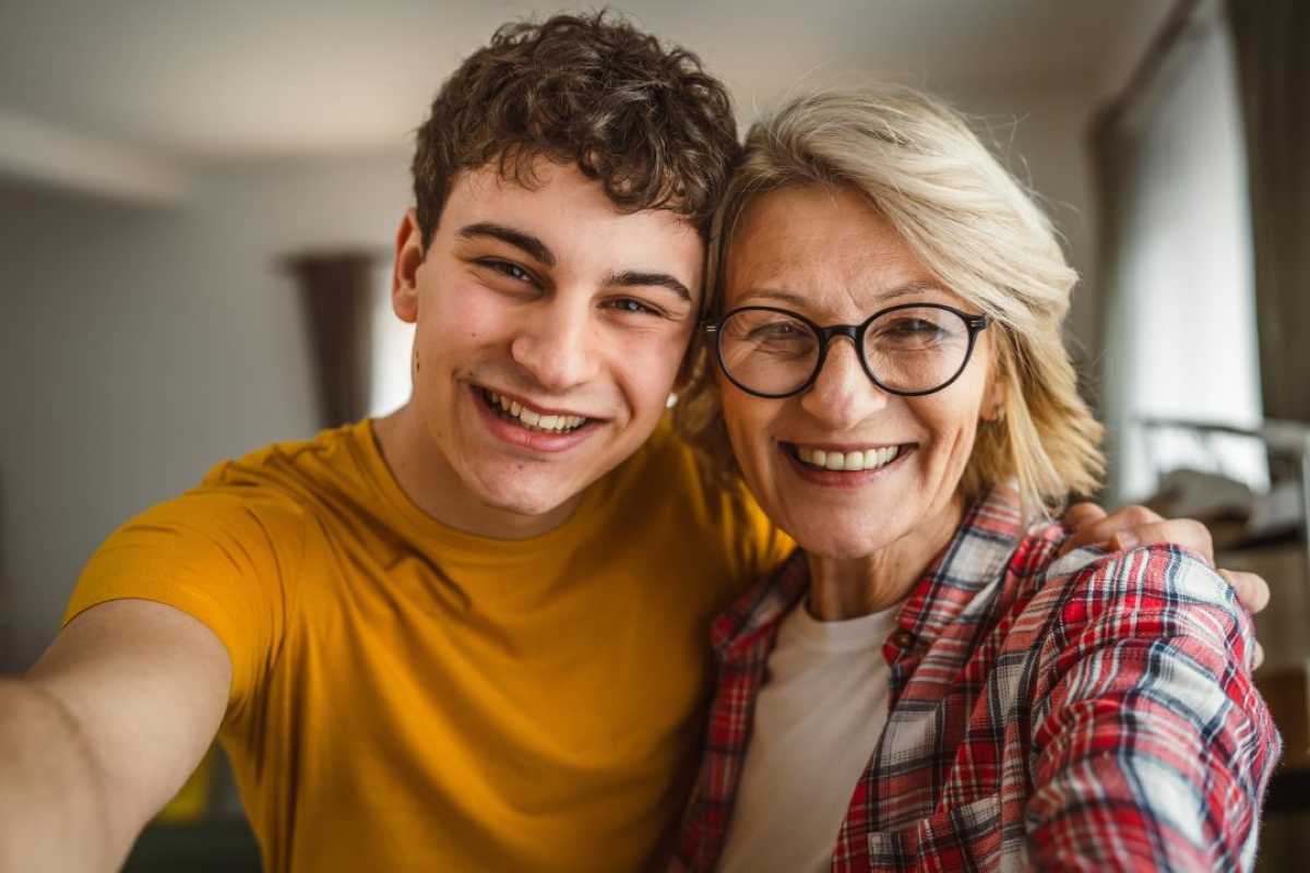 Smiling teenage boy and older woman taking a cheerful selfie together indoors, with their faces close.