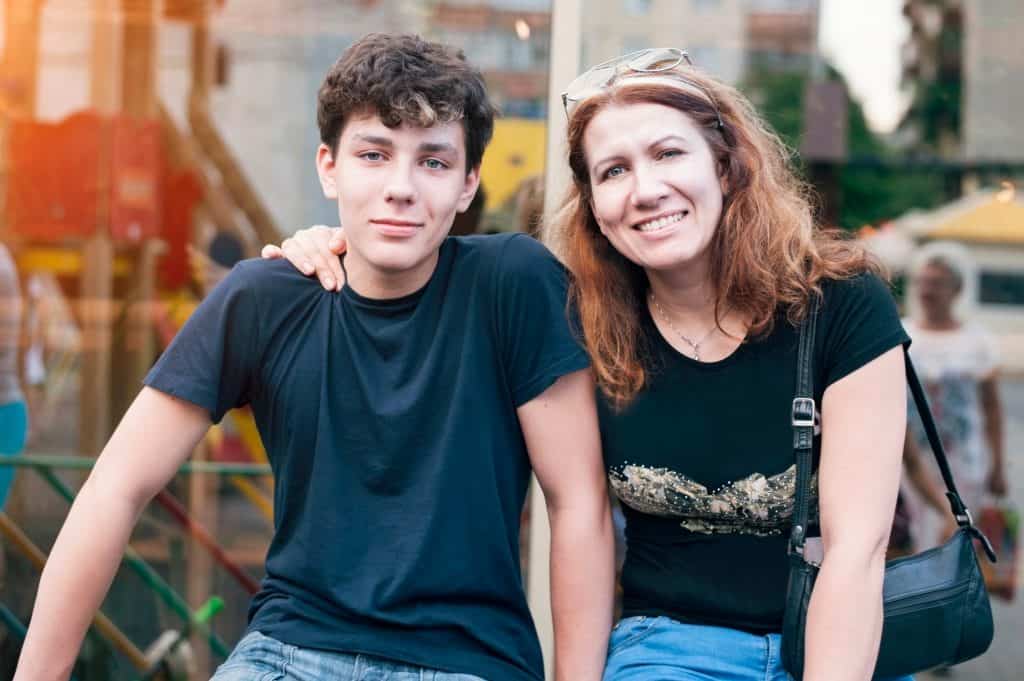 Teenage boy and woman posing outdoors with smiles, her hand resting on his shoulder in a casual, friendly gesture.