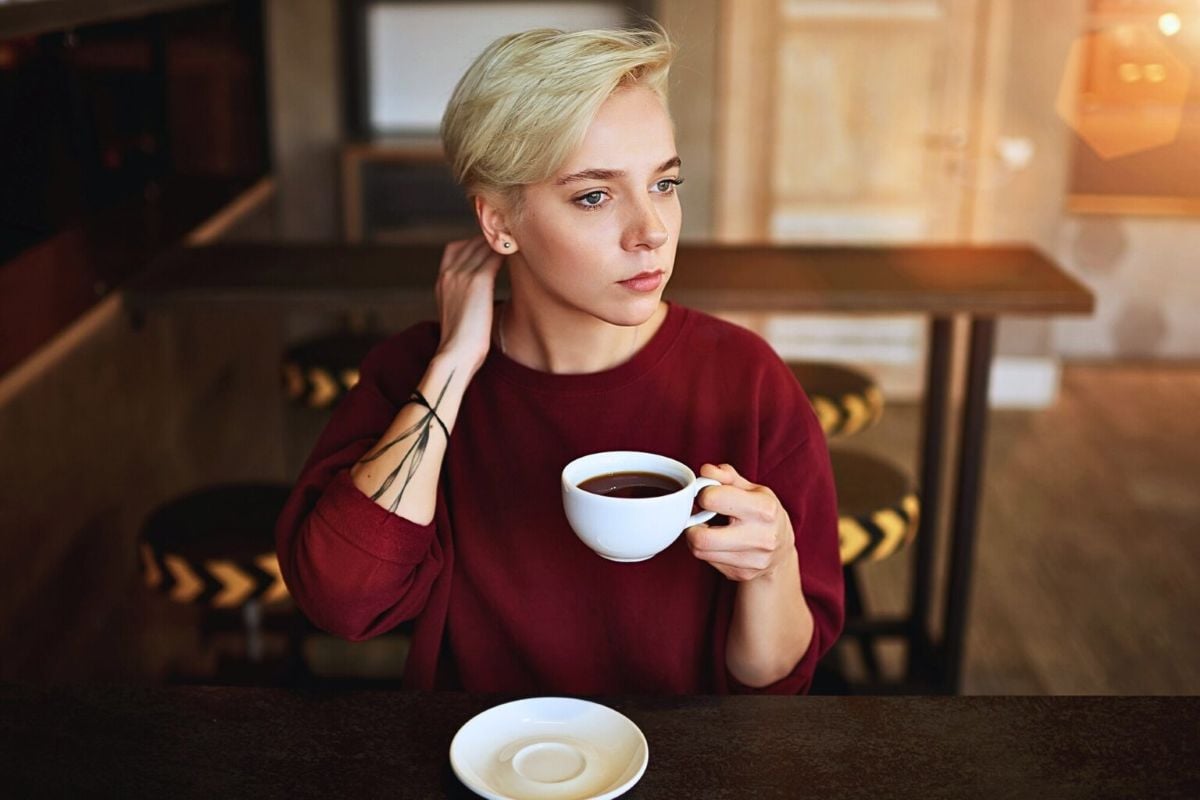Young woman with short blonde hair holding a coffee cup, gazing thoughtfully while sitting alone in a cozy café.