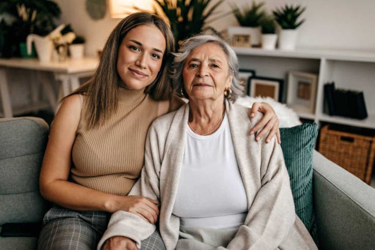 Young woman and elderly grandmother sitting closely on a couch, smiling gently in a cozy, plant-filled living room.