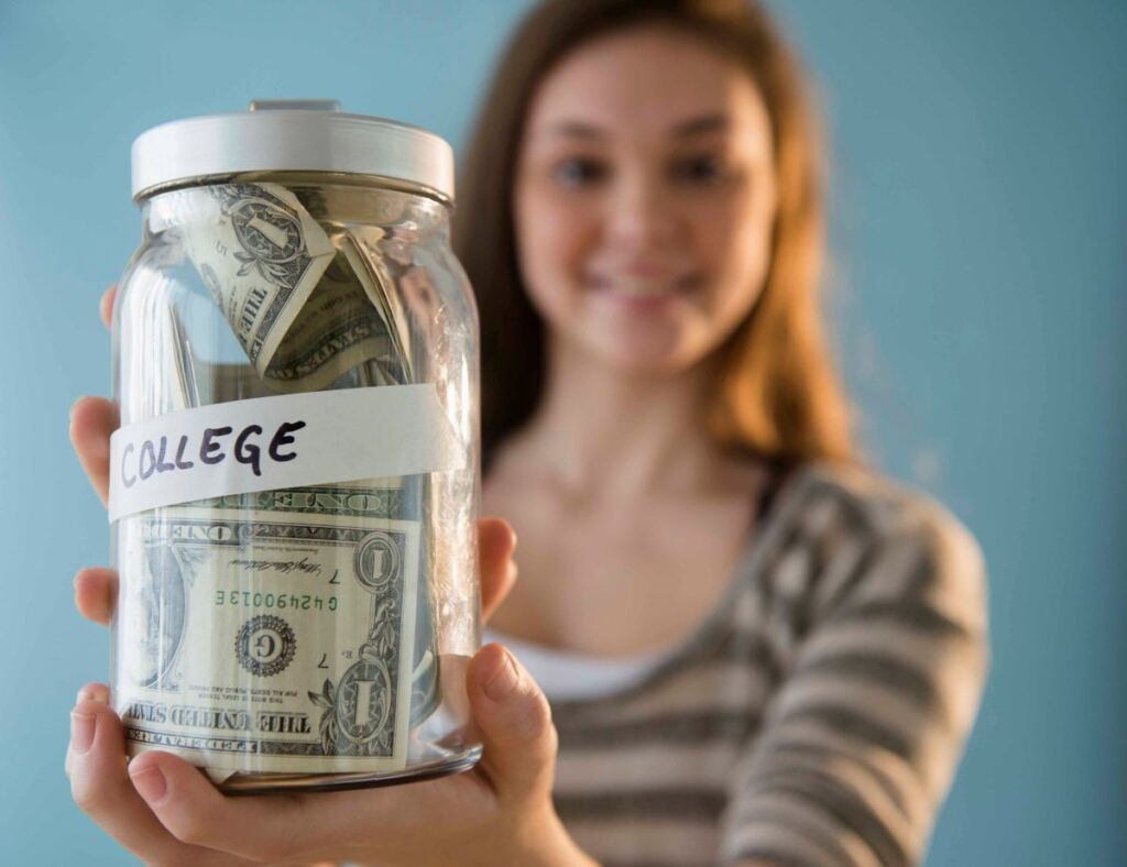 A person holds a glass jar labeled "COLLEGE," filled with rolled-up dollar bills, symbolizing savings for higher education.