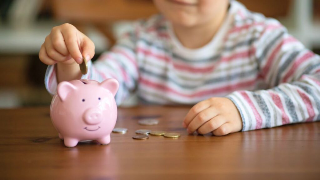 The Beginner’s Guide on How to Save Money for Your Grandchildren 4 A child in a striped shirt inserts a coin into a pink piggy bank, surrounded by additional coins on a wooden table.