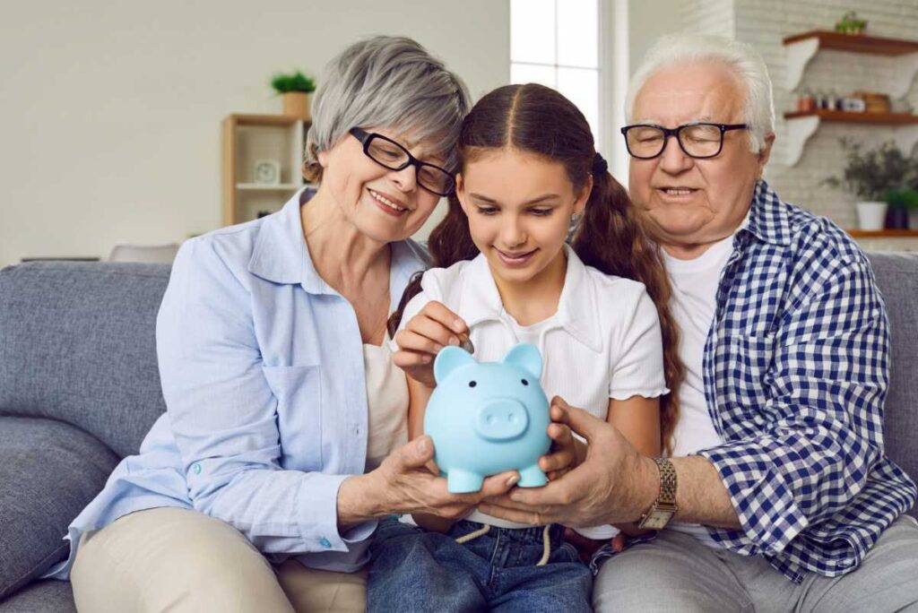 The Beginner’s Guide on How to Save Money for Your Grandchildren 3 An elderly couple and a child collaboratively hold a blue piggy bank, sharing moments of financial learning and family bonding on a sofa.