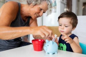 An adult and a child together at a table, with the adult inserting money into a blue piggy bank and a small red pot nearby.