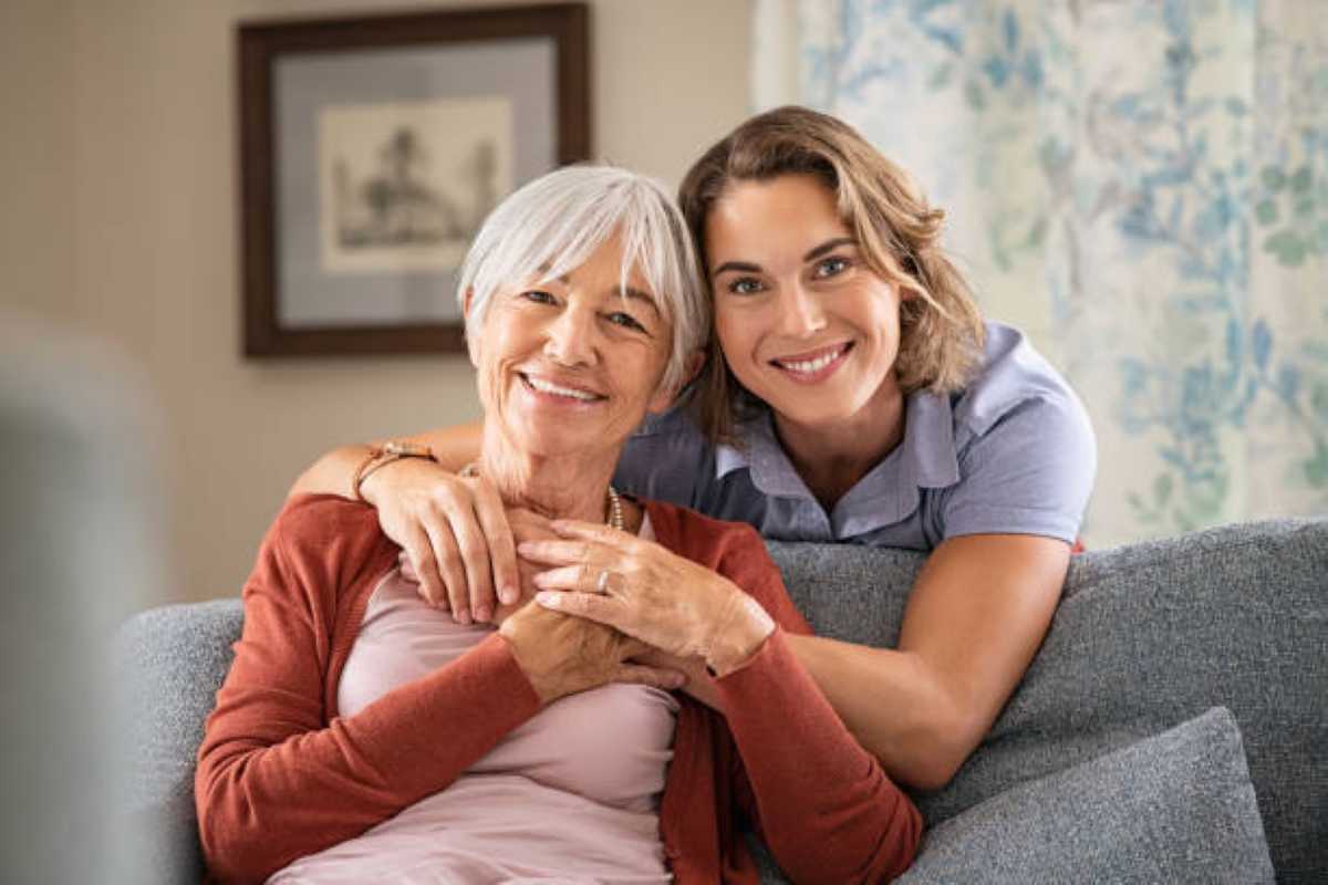 A woman in a cardigan embraces another seated on a couch, conveying warmth and affection in a cozy home setting.