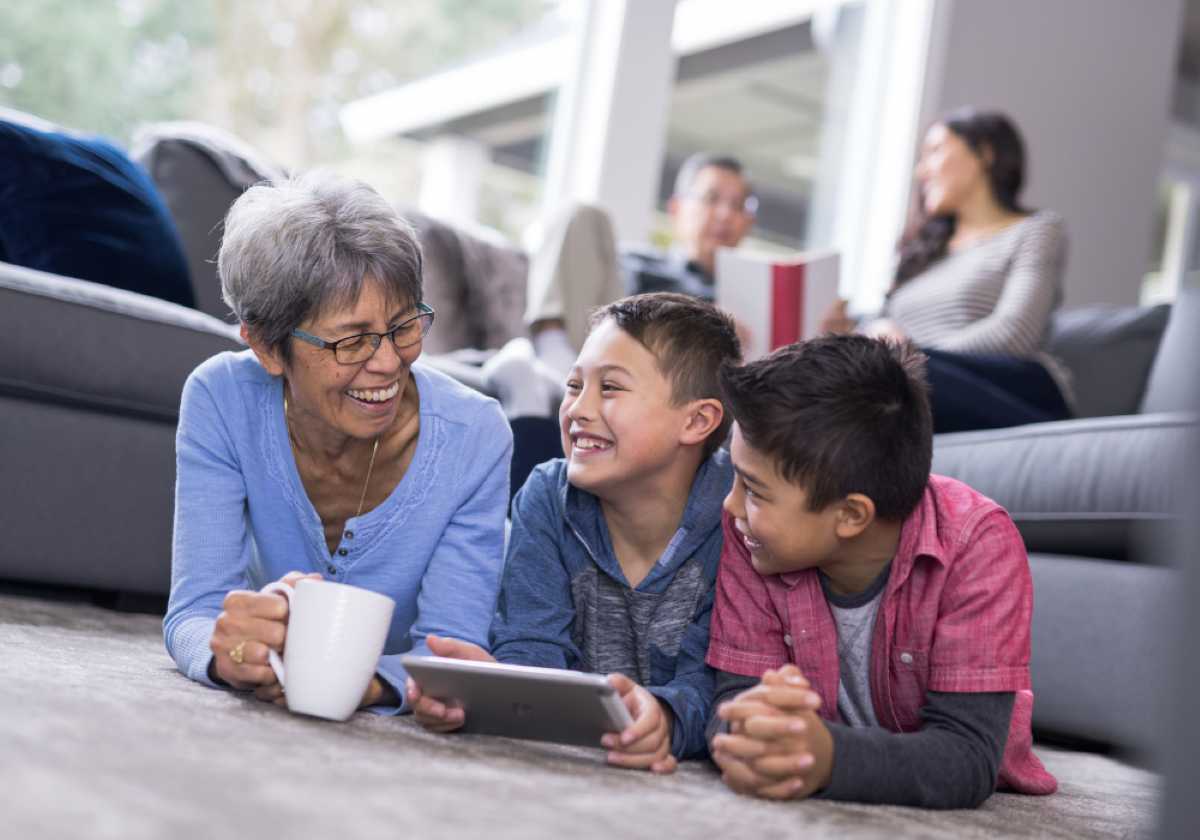 Smiling grandmother and two boys lying on the floor, enjoying a tablet together in a cozy living room.