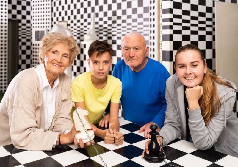 Two grandparents and two kids posing at a checkered table in a puzzle room, holding large chess pieces and game props.