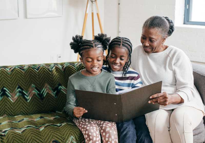 Grandmother reading a photo album with two young girls on a couch, all smiling and enjoying the moment.