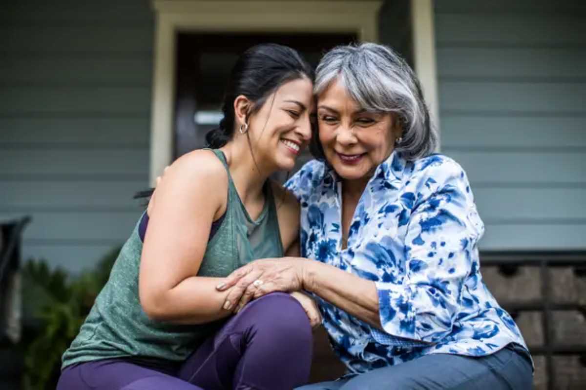 Smiling older woman and younger woman sit close together on a porch, holding hands and sharing a warm, affectionate moment.