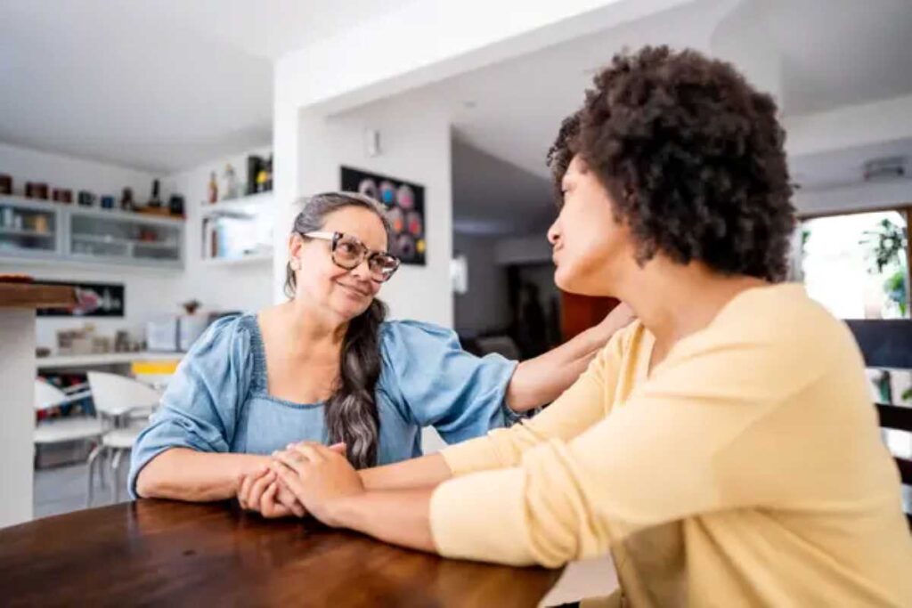 These are the 6 phrases adult children still want to hear from their parents, experts agree 3 Older woman gently holds a younger woman’s hands at a table, offering comfort and support in a heartfelt conversation.