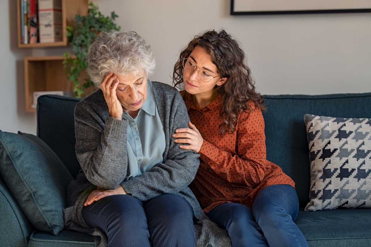 A woman comforts an elderly person sitting on a sofa, conveying warmth and support in a cozy living room setting.