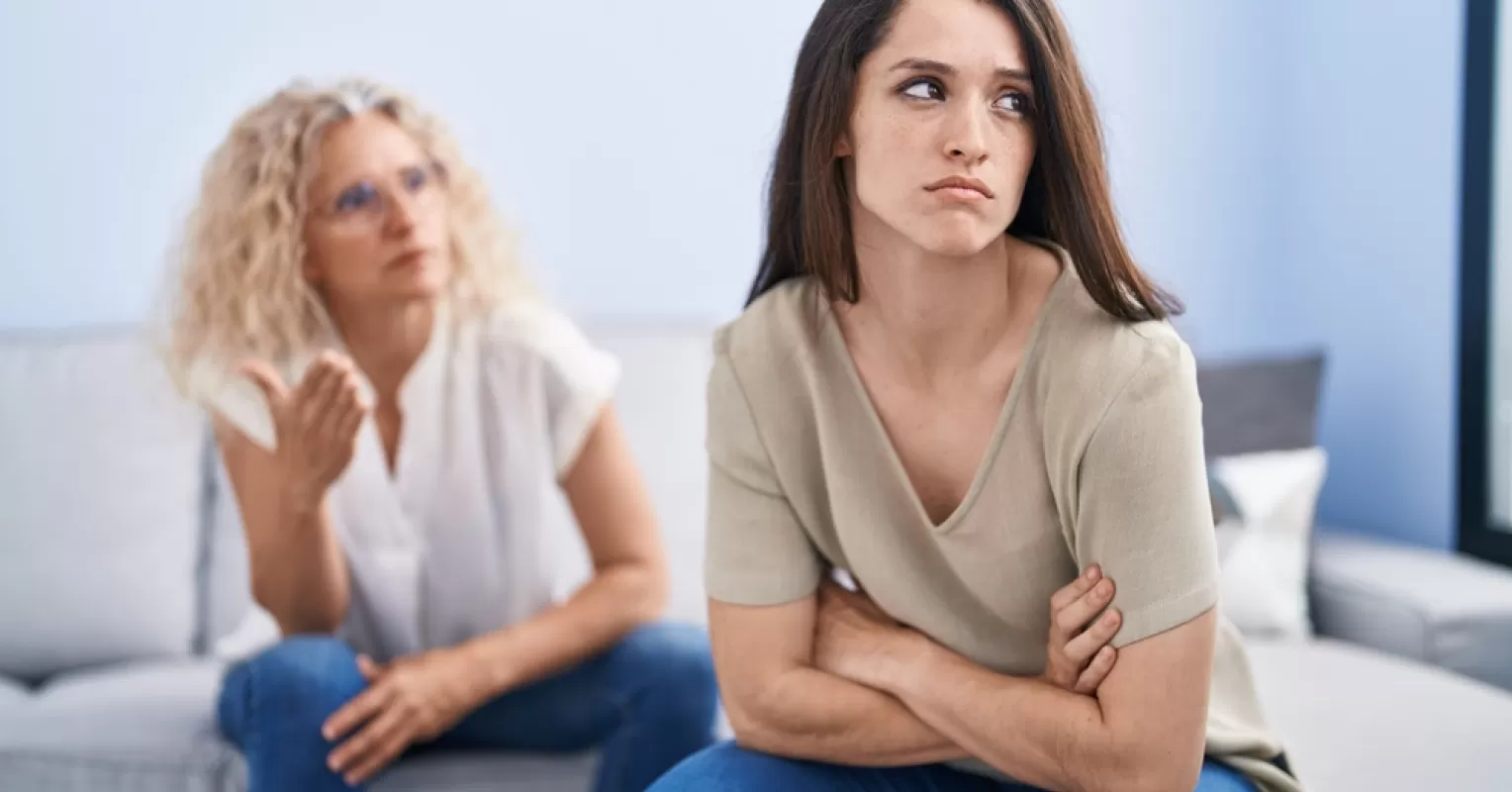 A young woman looks away, arms crossed, as an older woman gestures during a tense conversation in a modern living room.