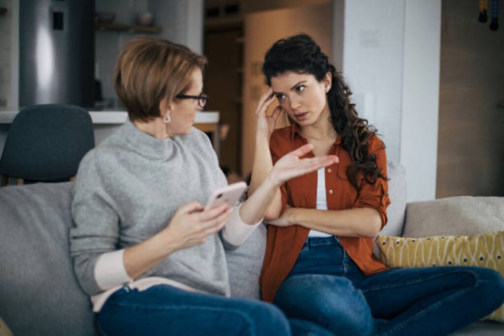11 Ways Parents Unknowingly Destroy Their Relationship With Their Grown Children 2 Two women sit on a couch having a tense conversation; the older woman gestures while the younger looks frustrated and touches her forehead.