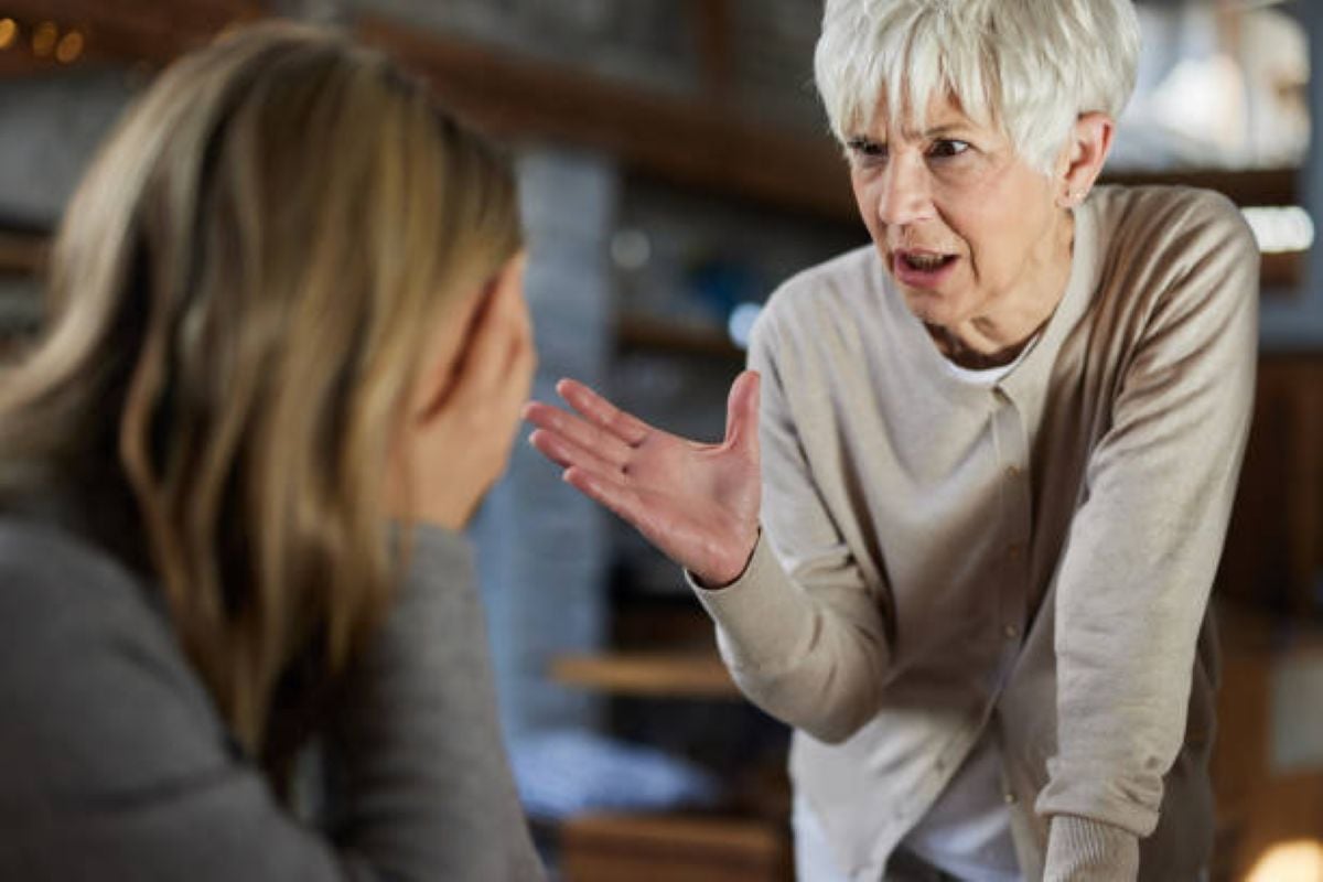 Older woman angrily gestures at a younger woman who sits with her face in her hands, suggesting a heated argument or confrontation.