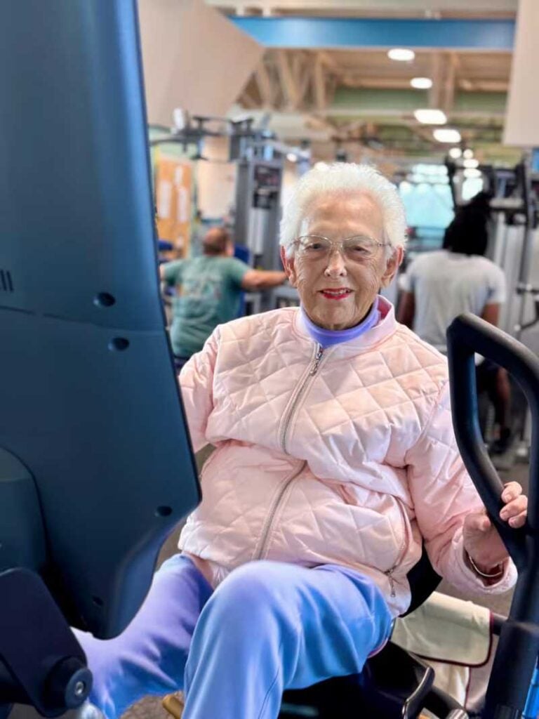 Elderly woman in pink jacket smiling while using a recumbent bike at a fitness center.