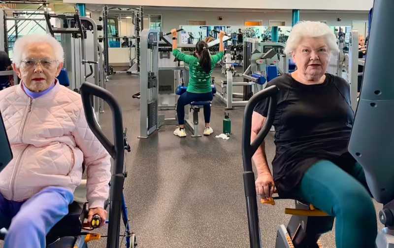 Two elderly women exercising on stationary bikes in a gym, with a younger woman working out in the background.