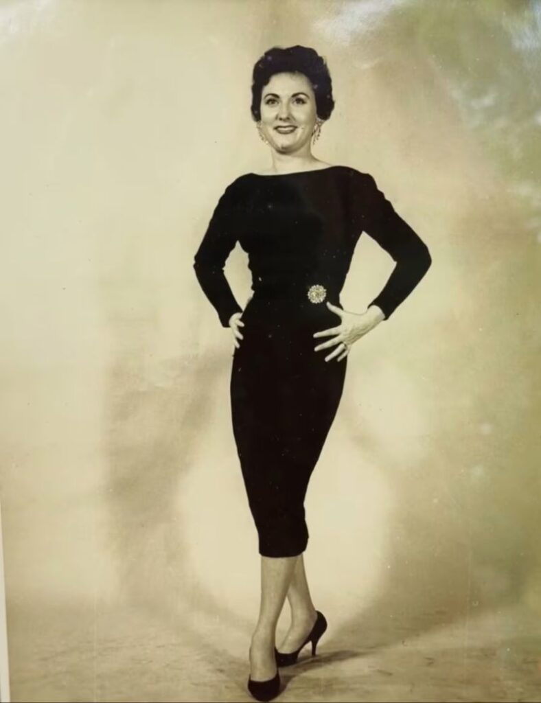 Vintage full-body portrait of a woman in a black dress and heels, posing confidently with hands on her waist.