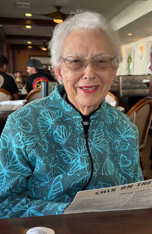 Smiling elderly woman in a turquoise butterfly-print jacket reading a menu at a restaurant.