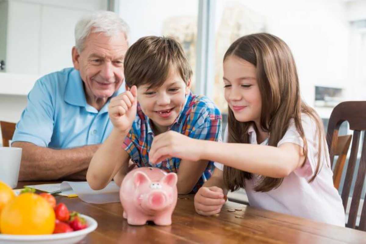 A smiling grandfather sits at a wooden table watching his two grandchildren—a young boy and girl—drop coins into a pink piggy bank. The children look happy and focused as they save money together, with a bowl of fresh fruit nearby on the table.