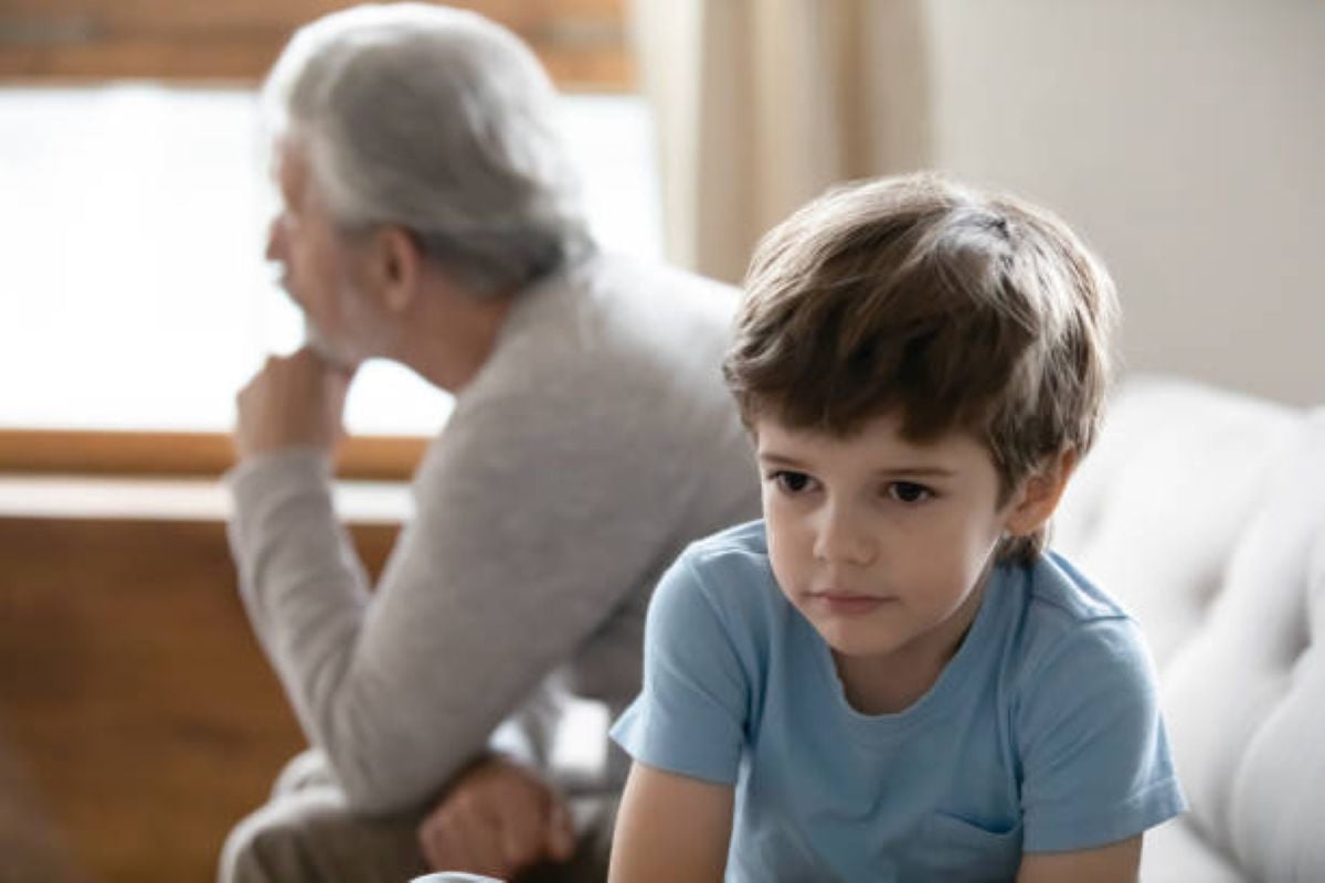 Image Alt Text Generator said: Sad-looking young boy in a blue shirt sits on a couch while an older man in the background looks away thoughtfully, both appearing distant or upset.