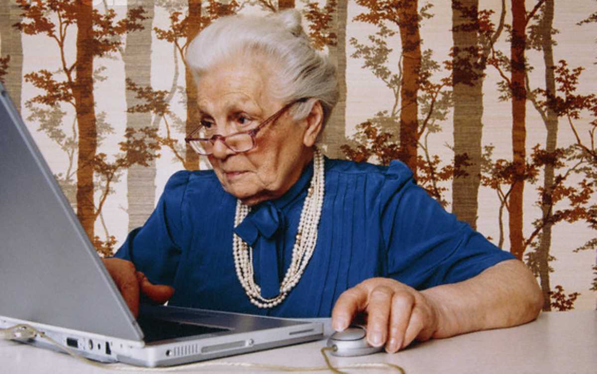 An elderly woman in a blue blouse sits at a table using a laptop, with hands on the keyboard and a mouse nearby, against a patterned background.