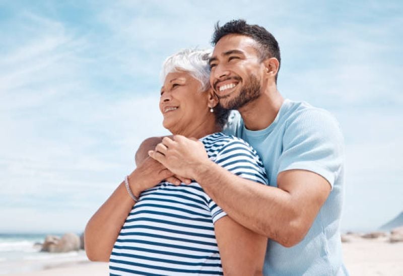 Smiling man hugging his elderly mother from behind as they enjoy a sunny day at the beach.