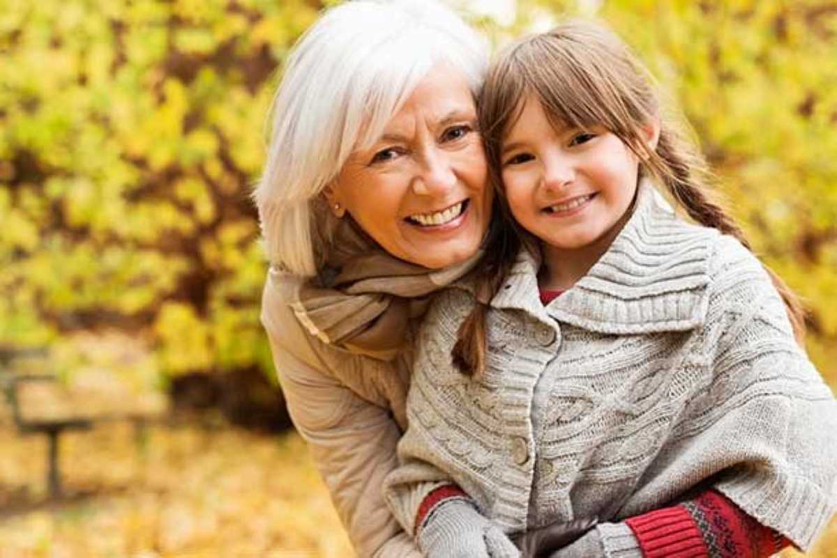 A smiling older woman with short white hair hugs a young girl outdoors in an autumn park, both wearing cozy sweaters as they pose closely together among yellow fall leaves.