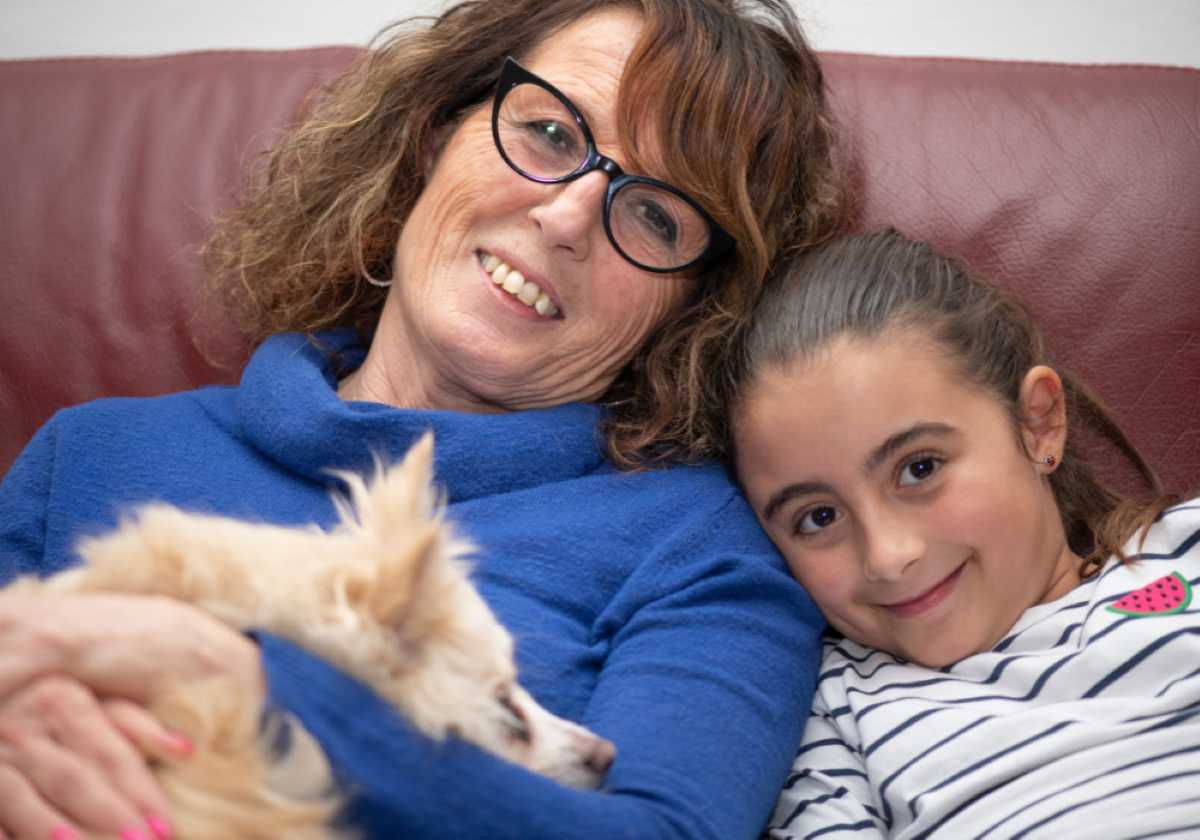 Smiling woman cuddling a small dog with a young girl resting her head on her shoulder, both relaxing on a red couch.