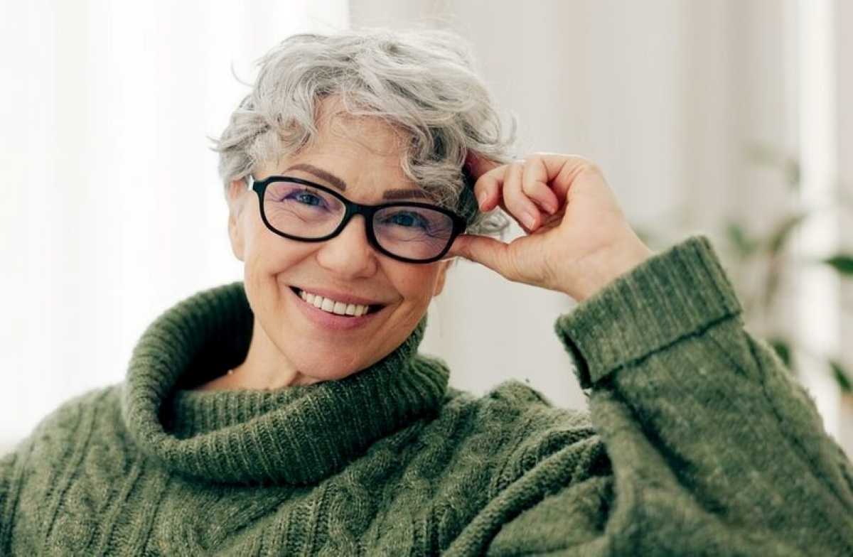 An elderly person with gray hair is seated, wearing a cozy green sweater, resting their hand on their head in a thoughtful pose.