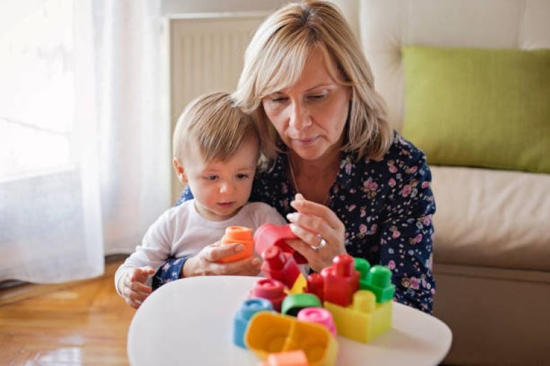 Grandmother and toddler playing with colorful plastic blocks at a small table indoors.