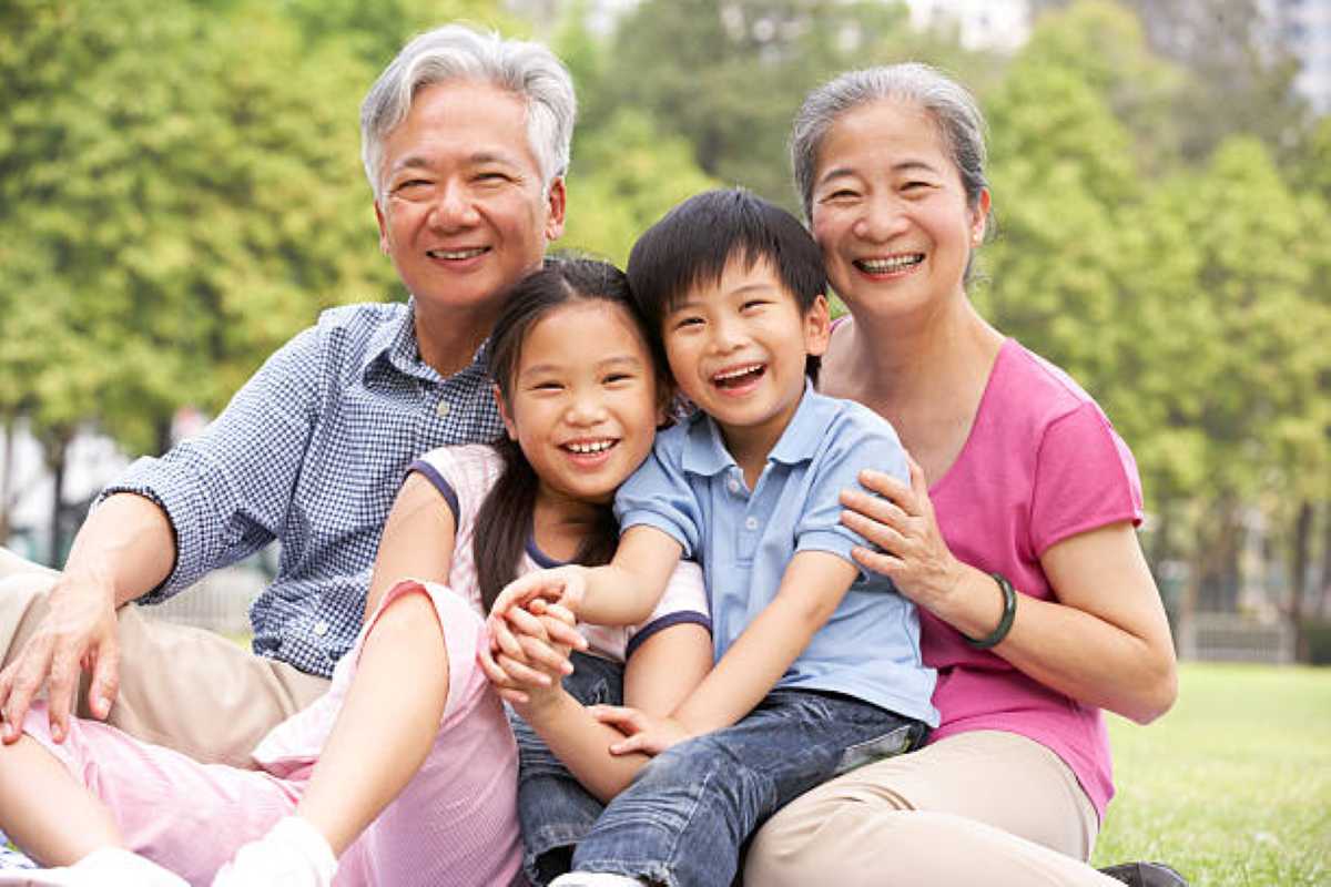 Smiling grandparents sitting on the grass and hugging their two cheerful grandchildren in a sunny park.