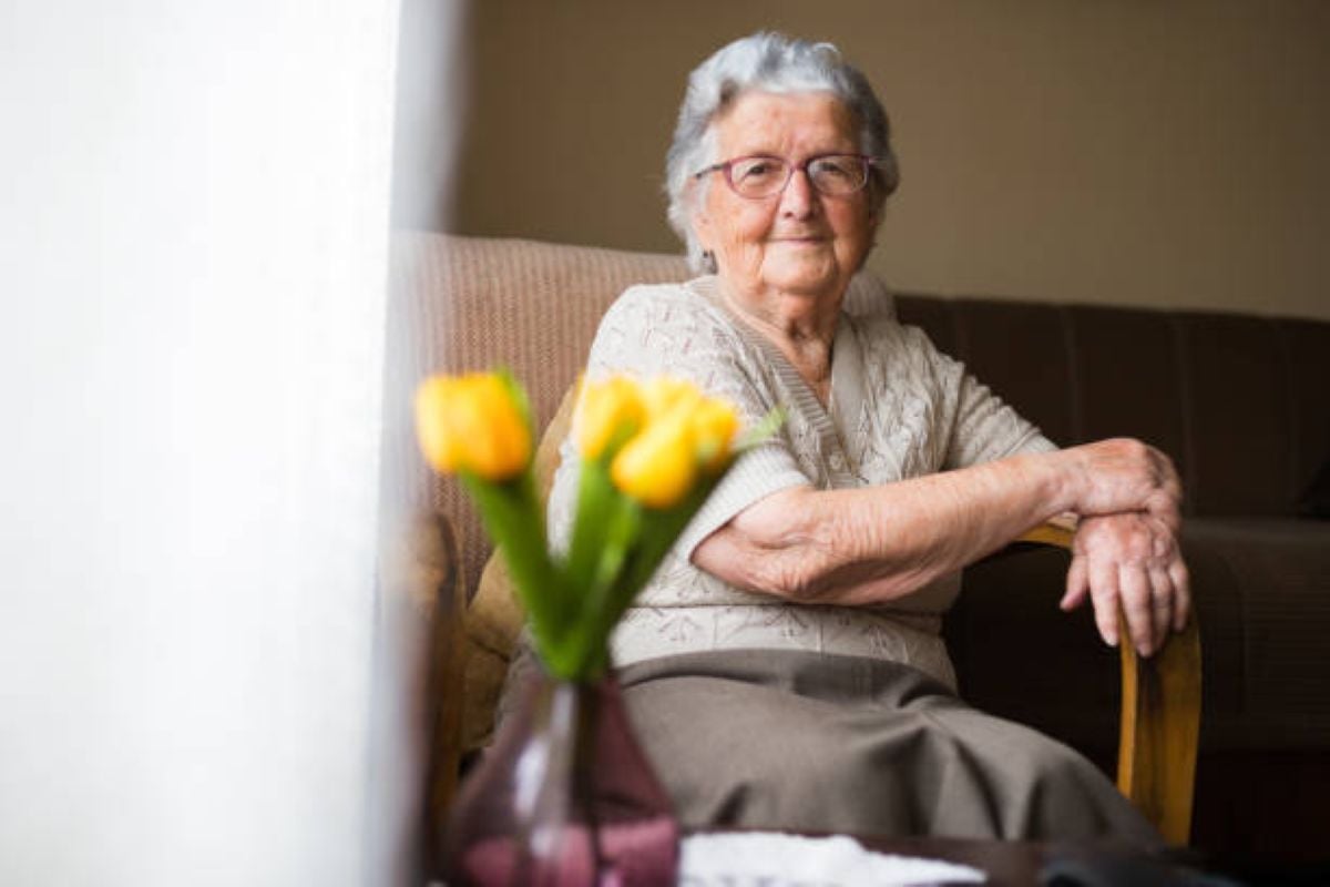 Elderly woman with gray hair and glasses sitting calmly on a couch near a window with yellow tulips in the foreground.