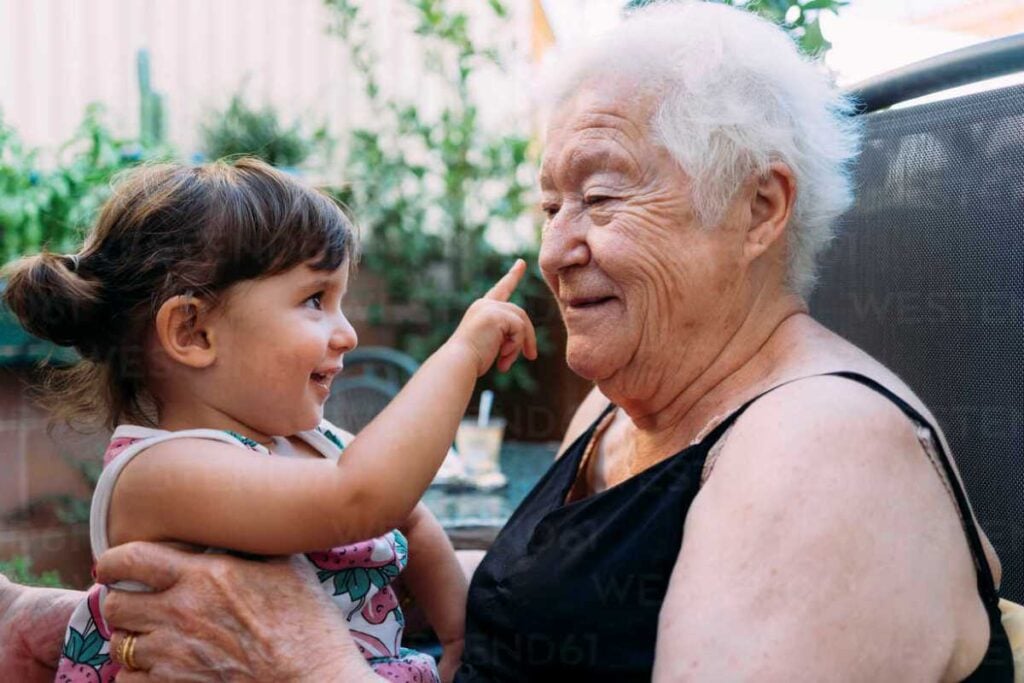 Laughing toddler touching elderly woman’s nose while sitting on her lap outdoors.