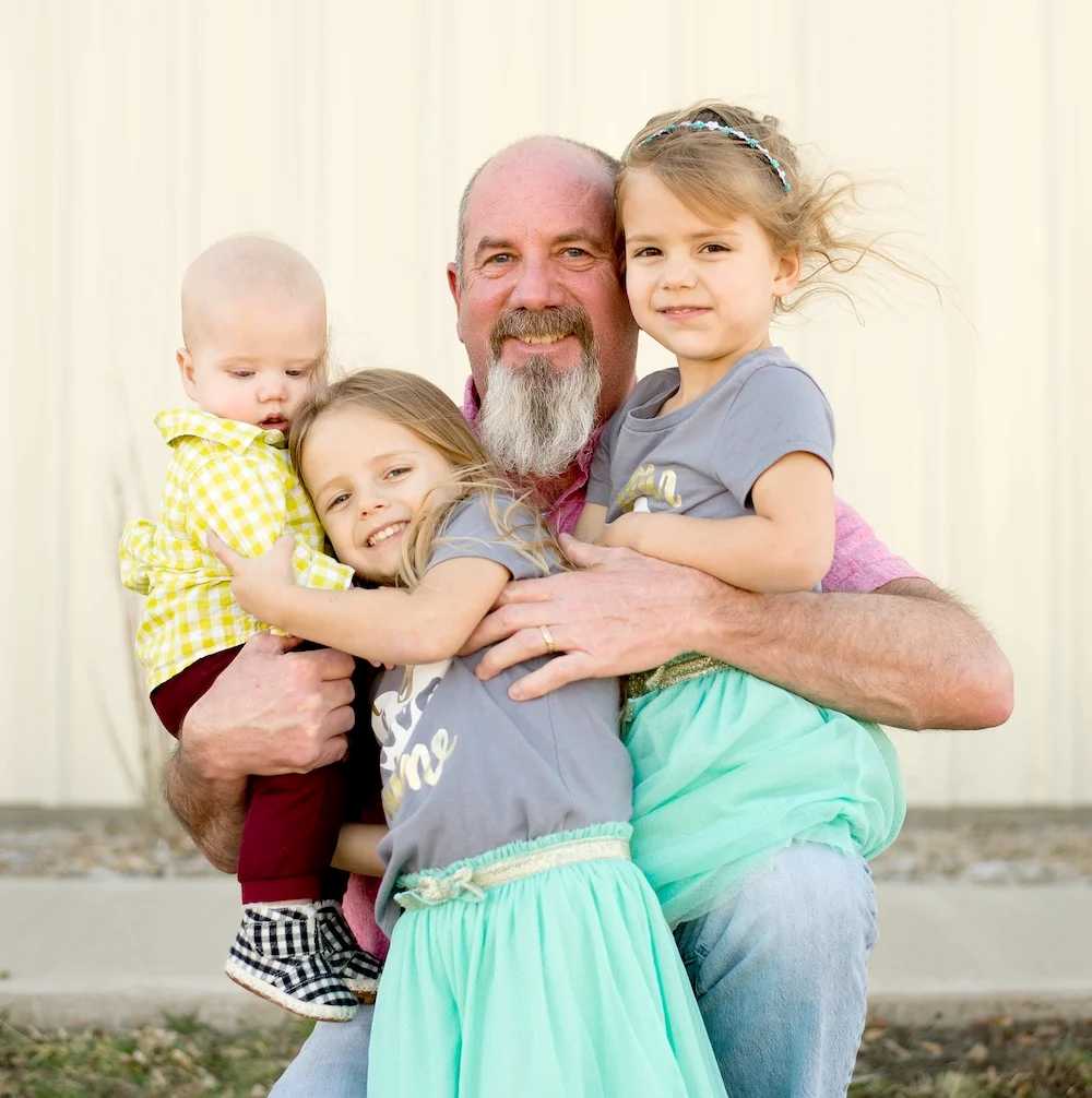 Bearded grandfather hugging three young grandchildren, smiling warmly outside.