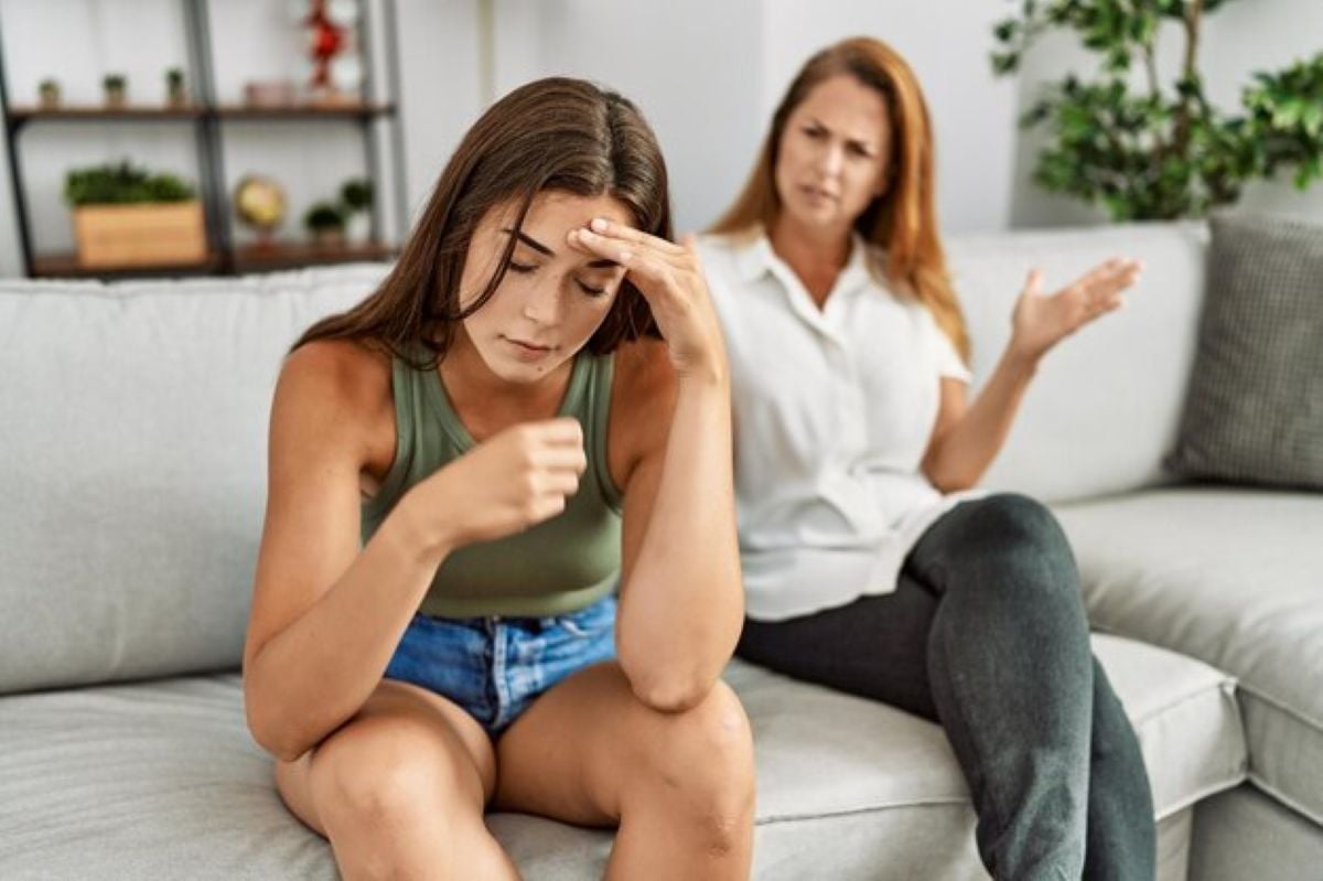 Two women sit on a light gray sofa, one with her head down, looking distressed, while the other gestures, appearing to engage in conversation.