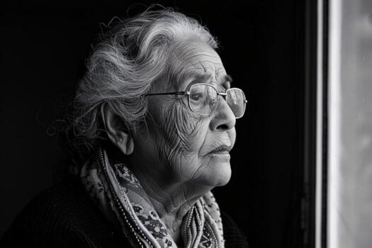 Elderly woman with glasses and a scarf gazing out a window in a black and white portrait, with a thoughtful expression.