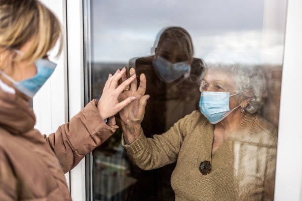 Elderly woman wearing a mask touching hands through a glass window with a younger woman during a pandemic visit.