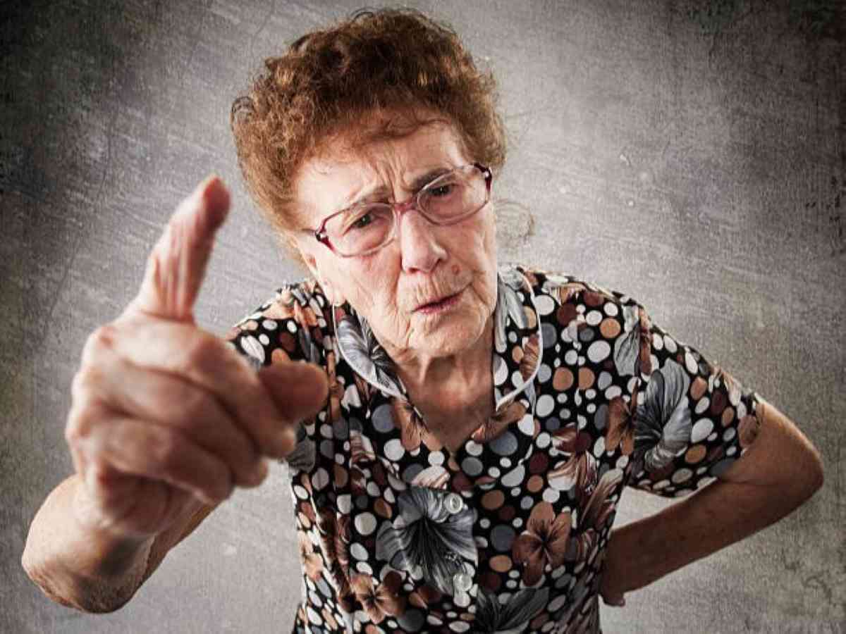 Elderly woman with glasses and a patterned shirt pointing her finger with a stern expression, as if scolding or warning someone.