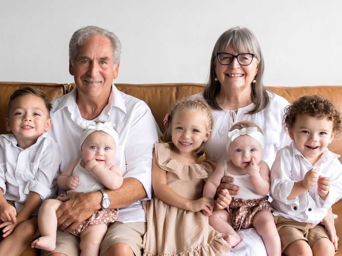 Smiling grandparents sitting on a couch with six young grandchildren, including two baby twins in matching outfits.