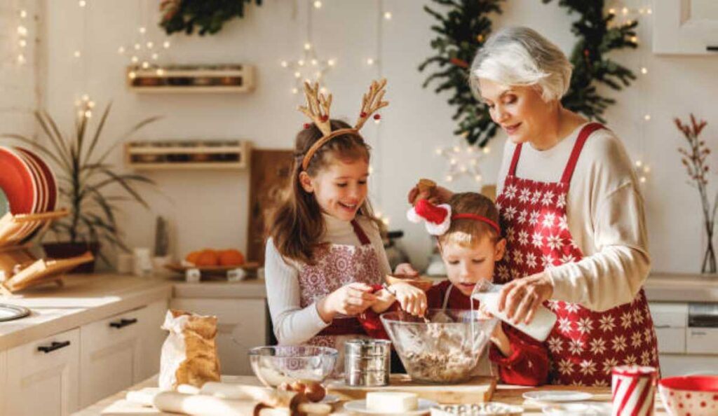 The 8 Christmas Grandma Personalities — Which One Do Your Grandkids See? 3 Smiling grandma wearing a red apron bakes Christmas treats with two young grandchildren in a cozy, decorated kitchen filled with holiday lights.