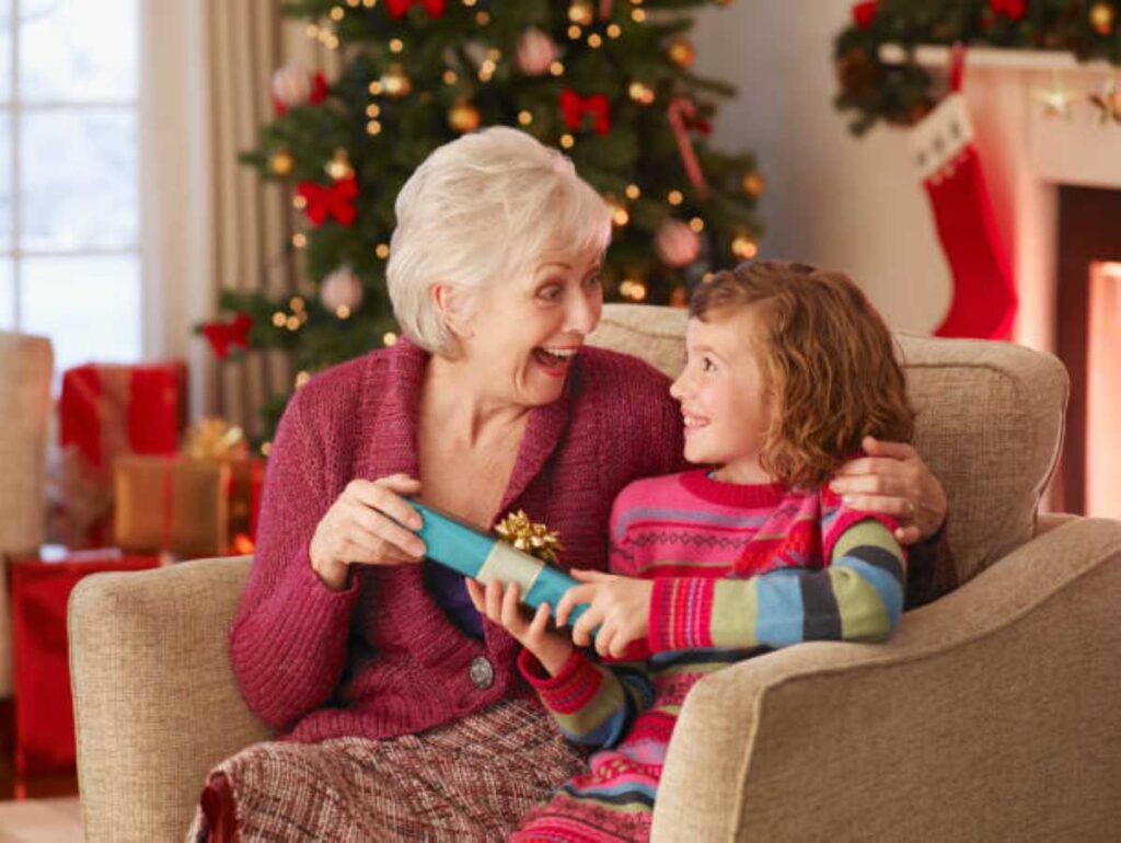 The 8 Christmas Grandma Personalities — Which One Do Your Grandkids See? 2 Grandma and young grandchild sit together on a couch opening a Christmas gift, smiling at each other in a cozy living room with a decorated Christmas tree in the background.
