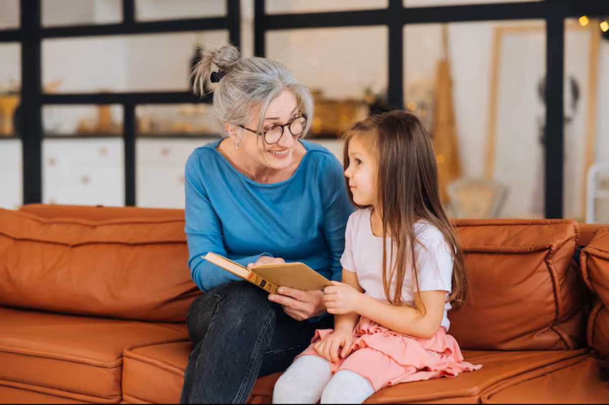 Grandmother sitting on a couch reading a book to her young granddaughter, smiling and talking gently as they share a calm, attentive moment together.