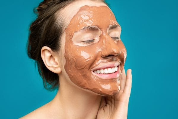 36 Unexpected Habits That Show You’re Actually on Track for a 100-Year Life 6 A young woman with her eyes closed smiles while wearing a brown clay face mask against a bright teal background.