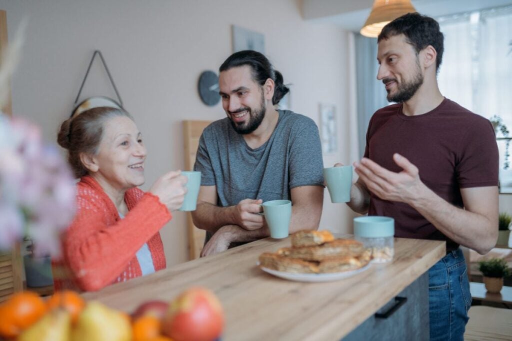 Parents who maintain strong bonds with their adult children exhibit these 6 behaviors 2 Elderly woman and two men smiling and chatting over coffee at a kitchen counter with a plate of pastries in front of them.
