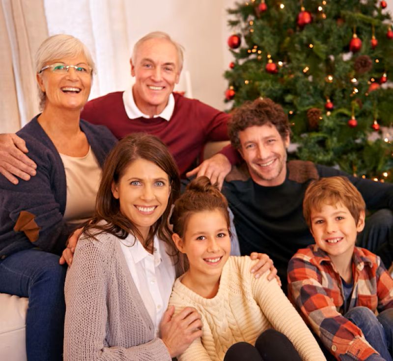 25 Simple Christmas Traditions You Can Start This Year That Grandkids Never Forget 3 A family gathers on a couch, smiling and posing together in front of a decorated Christmas tree with red ornaments.