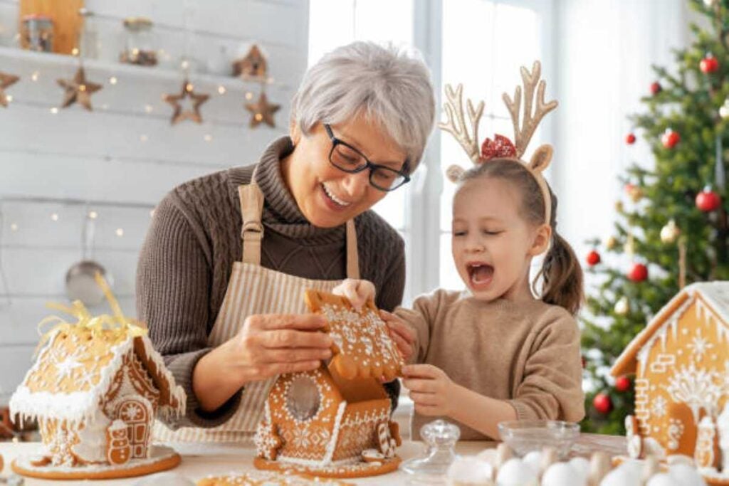 25 Simple Christmas Traditions You Can Start This Year That Grandkids Never Forget 4 Grandma and granddaughter decorate a gingerbread house together at Christmas, laughing and wearing reindeer headbands beside a festive holiday table.