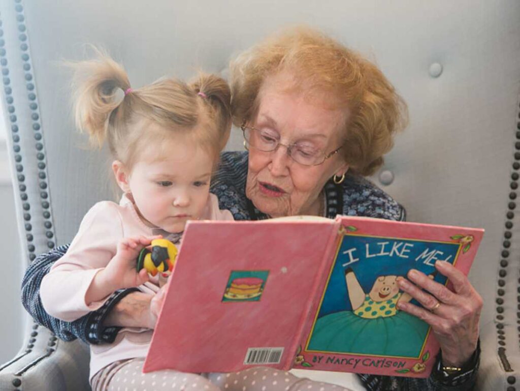 The Most Influential Grandmas Make a Massive Difference Without Overstepping Boundaries by Doing These 7 Things 2 An elderly woman sits in a cushioned chair reading a picture book titled “I Like Me!” to a young girl with pigtails. The toddler holds a small toy while listening closely, creating a warm and cozy moment together.