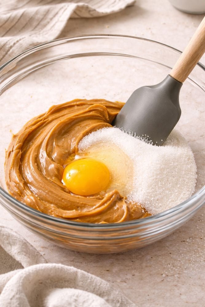 The Best 3-Ingredient Peanut Butter Cookies Recipe You’ll Ever Make 3 Glass mixing bowl with peanut butter, sugar, and a raw egg being combined with a spatula to make cookie dough.